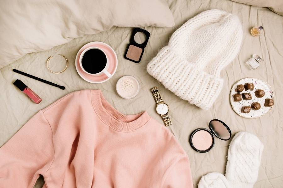 Spring fashion trends flat lay with pink sweater, white beanie, coffee cup and makeup essentials on cream bedding