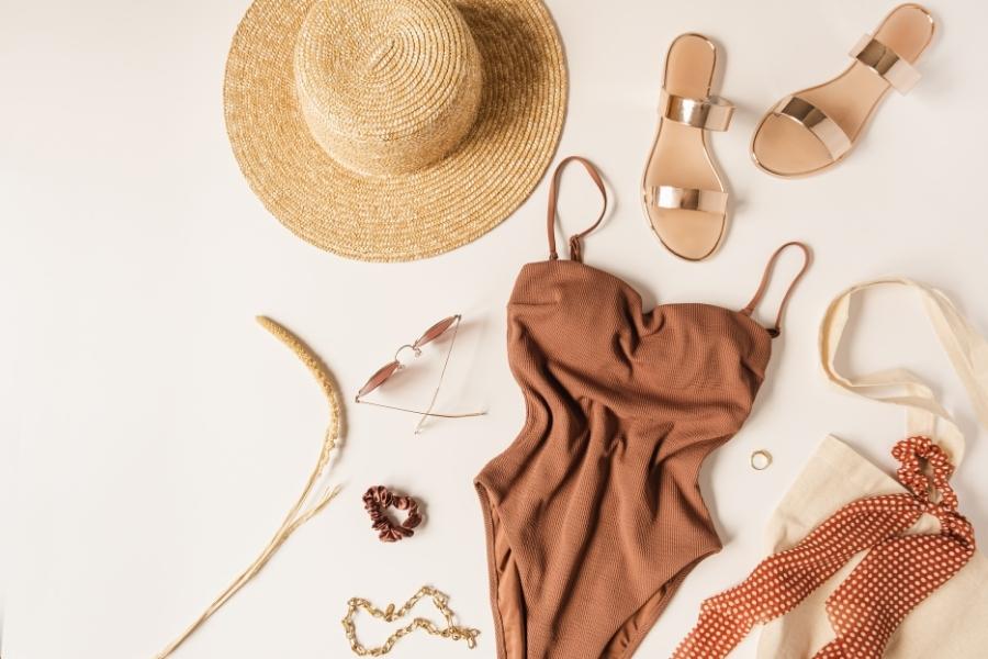Flat lay of trending accessories including straw hat, gold sandals, brown dress and jewelry on neutral background