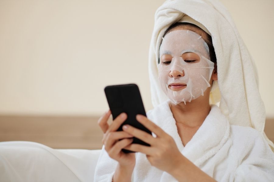 Woman enjoying relaxing skincare routine moment with face mask while using smartphone at home