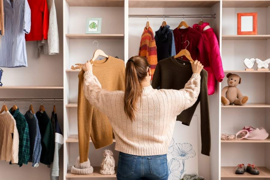 Woman organizing capsule wardrobe pieces in white closet with colorful sweaters and accessories