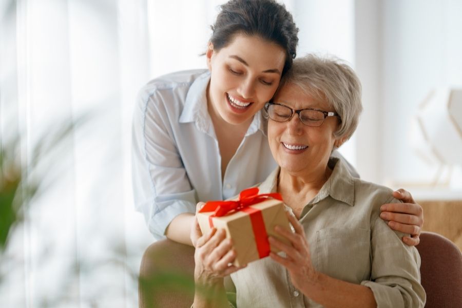 Daughter giving wrapped Mother's Day gift to smiling elderly mother in bright living room