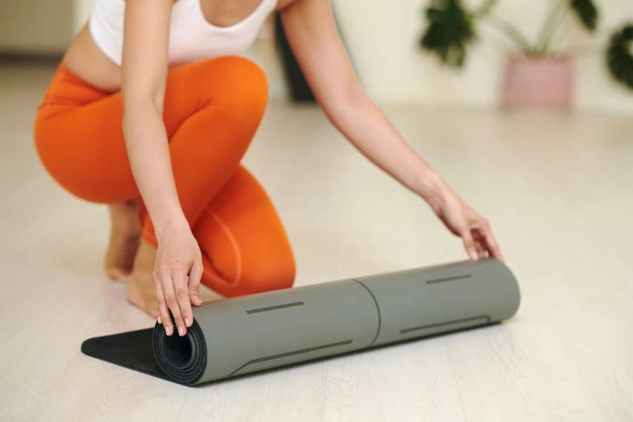 A woman wearing orange leggings rolling out a grey yoga mat on a bright wooden floor, starting her mindful morning routines with a peaceful exercise session.